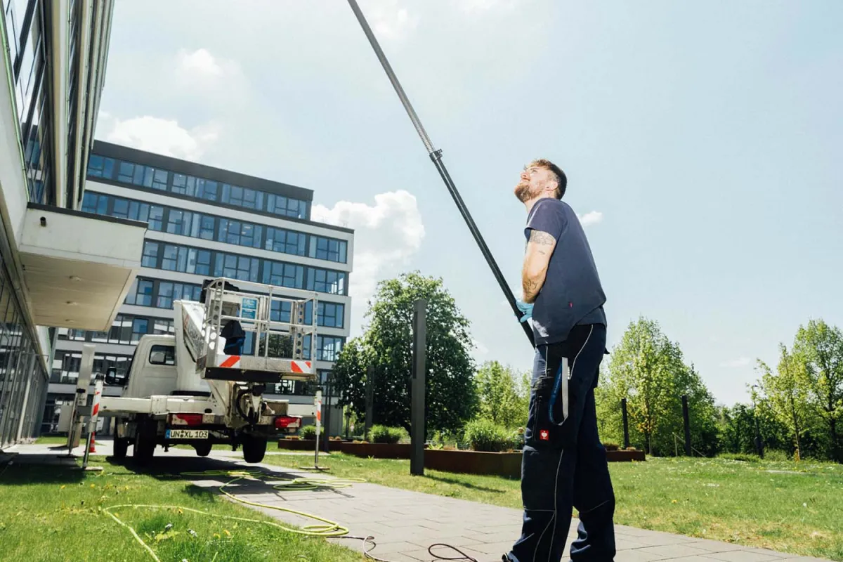 Mitarbeiter mit Teleskopstange vor einem Bürogebäude, dahinter ein Hubsteiger